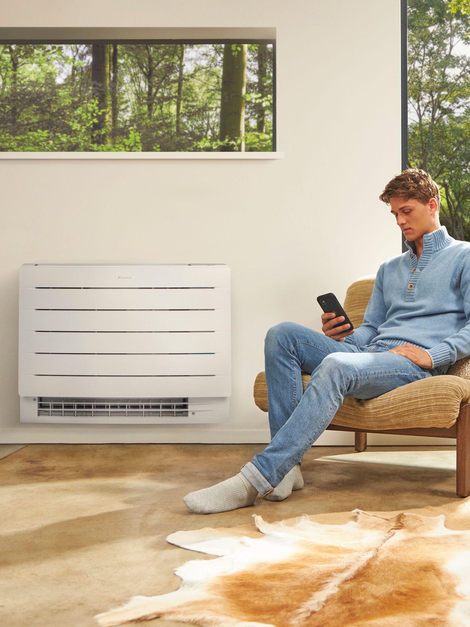 Muškarac sjedi u svijetloj prostoriji i gleda u svoj telefon s dizalicom topline ugrađenom na zid iza njega