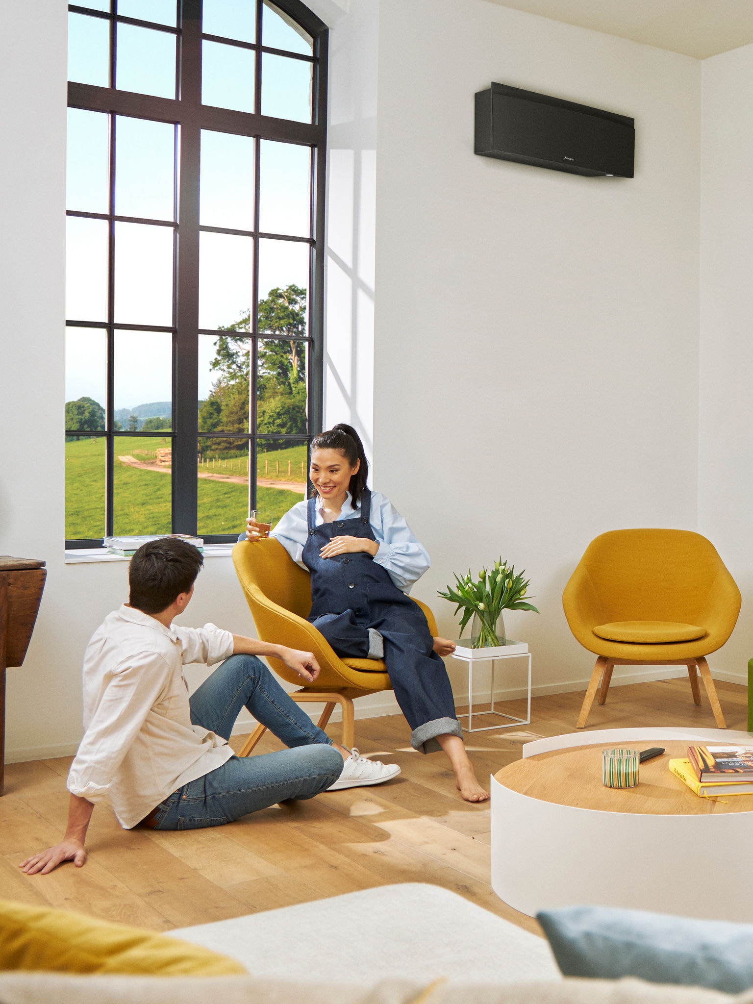 Muškarac i žena sjede ispred velikog prozora u svijetloj prostoriji s crnom zidnom dizalicom topline ugrađenom na zid iza njih