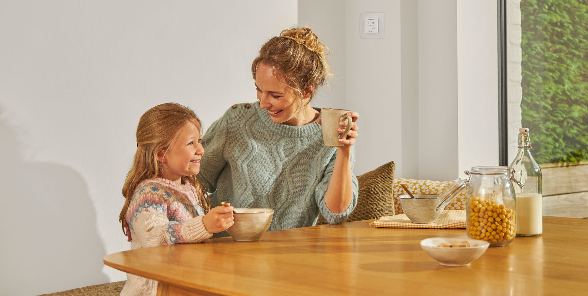 Djevojka koja jede iz zdjelice i sjedi za stolom pokraj žene koja u ruci drži šalicu, obje se smiješe, sretne su i opuštene te uživaju u zdravom zraku.